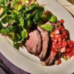 Grilled tri-tip sliced against the grain on a wooden cutting board, showing a smoky charred crust and pink juicy interior, garnished with fresh herbs