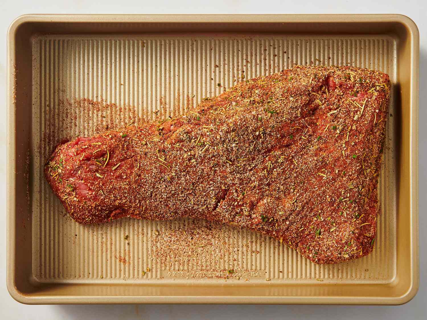 Tri-tip roast coated in smoky dry rub on a sheet pan next to small bowls of spices including smoked paprika, garlic powder, and black pepper