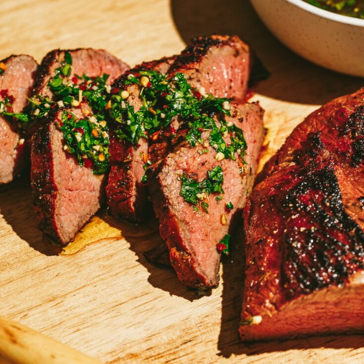 Close-up of thinly sliced grilled tri-tip fanned on a platter with a ramekin of chimichurri sauce and grilled corn in the background