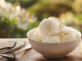 Homemade vanilla ice cream scooped into a white ceramic bowl, golden and creamy with visible vanilla bean specks, styled on a rustic wooden table with a linen napkin and fresh vanilla pods