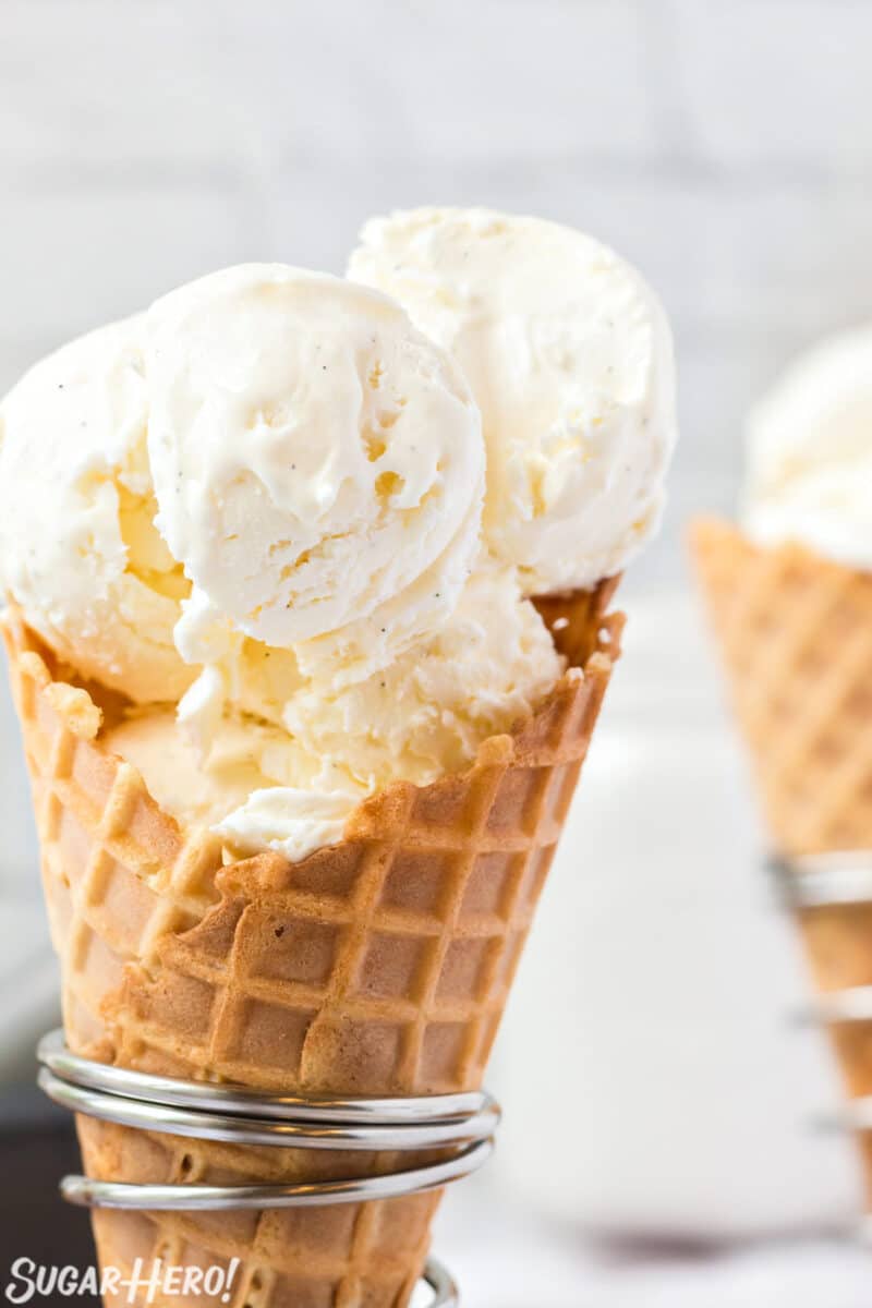 Close-up of homemade vanilla ice cream scooped into a waffle cone, showing creamy pale golden texture with black vanilla bean specks, soft focus background of a sunny outdoor summer setting