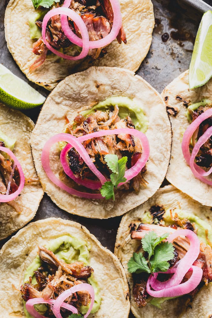 Close-up of crispy carnitas taco on a corn tortilla with charred edges, pickled red onions, cilantro, and a lime wedge on a dark slate surface