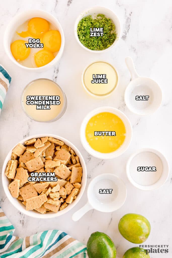 Key Lime Pie ingredients flat lay with Key limes, condensed milk, graham crackers, eggs, and butter on a marble surface
