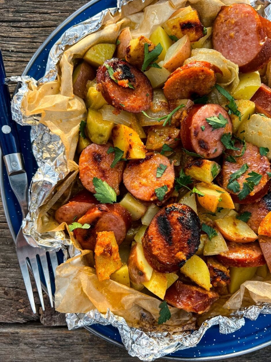 Close-up of opened kielbasa and potato foil packet showing caramelized sausage slices, tender golden potatoes glistening with butter and spicy mustard sauce, steam rising