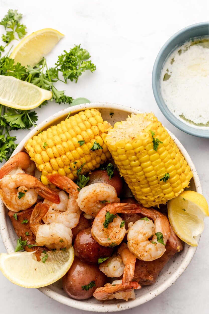 Overhead flat-lay of raw shrimp, andouille sausage sliced on a cutting board, red potatoes, corn on the cob, Old Bay seasoning tin, lemons, and garlic on a rustic wooden surface