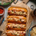 Classic Maine Lobster Roll served in a golden toasted split-top bun overflowing with creamy lobster salad, garnished with fresh chives, served on parchment with lemon wedges and kettle chips on a rustic wooden board outdoors