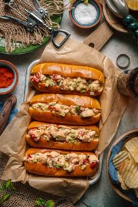 Classic Maine Lobster Roll served in a golden toasted split-top bun overflowing with creamy lobster salad, garnished with fresh chives, served on parchment with lemon wedges and kettle chips on a rustic wooden board outdoors