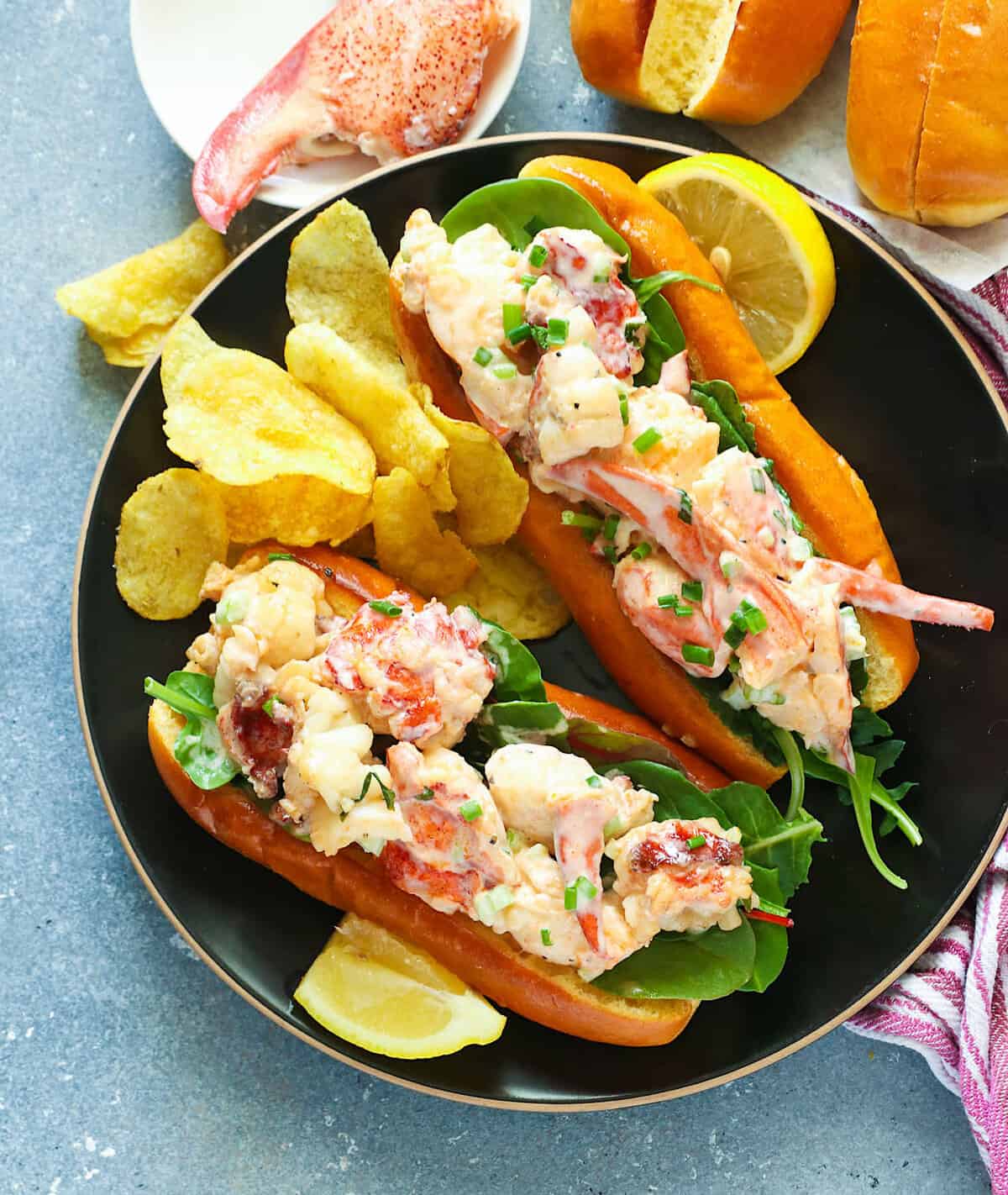 Step-by-step flat lay of Maine Lobster Roll ingredients — fresh cooked lobster claws and tails, mayo, lemon, chives, celery, and split-top buns on a marble surface with a prep bowl