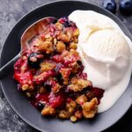 Mixed Berry Crisp with golden oat crumble topping served warm in a white baking dish with a scoop of melting vanilla ice cream, overhead shot on a rustic wooden table