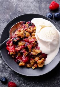 Mixed Berry Crisp with golden oat crumble topping served warm in a white baking dish with a scoop of melting vanilla ice cream, overhead shot on a rustic wooden table