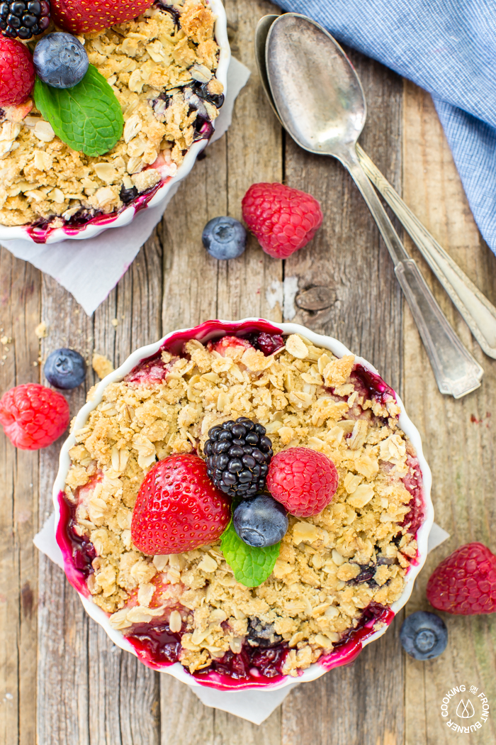 Overhead flat-lay of mixed berry crisp ingredients: fresh strawberries, blueberries, raspberries in separate bowls, oats, brown sugar, cold butter cubes, cinnamon, and lemon on a marble surface