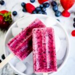 Mixed Berry Popsicles arranged on crushed ice with fresh strawberries, blueberries, and raspberries scattered around them, vibrant magenta and purple colors, bright natural lighting