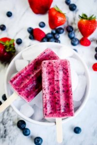 Mixed Berry Popsicles arranged on crushed ice with fresh strawberries, blueberries, and raspberries scattered around them, vibrant magenta and purple colors, bright natural lighting