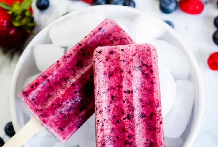 Mixed Berry Popsicles arranged on crushed ice with fresh strawberries, blueberries, and raspberries scattered around them, vibrant magenta and purple colors, bright natural lighting