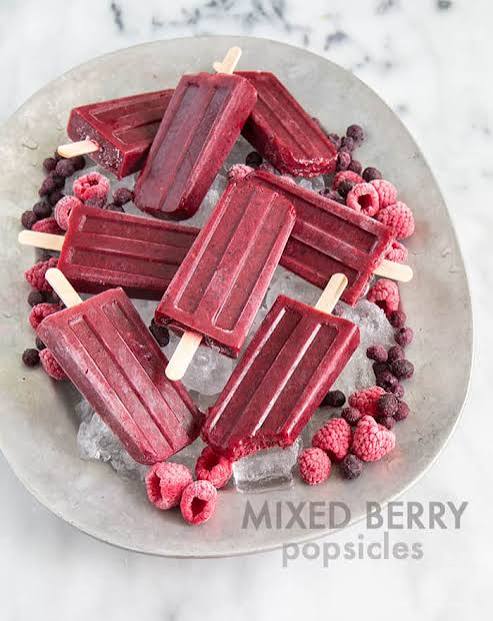 Close-up of a single Mixed Berry Popsicle being held against a bright summer background, showing the gorgeous jewel-toned color, whole berry pieces, and cream swirls in cross-section