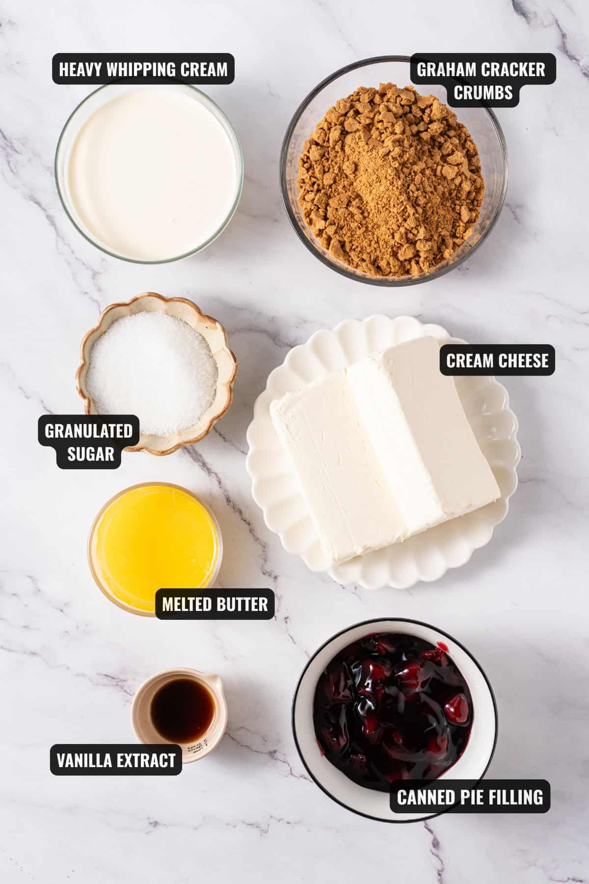 Overhead flat lay of no bake cheesecake ingredients — cream cheese blocks, graham crackers, heavy cream, lemon, vanilla extract, and butter on a marble countertop