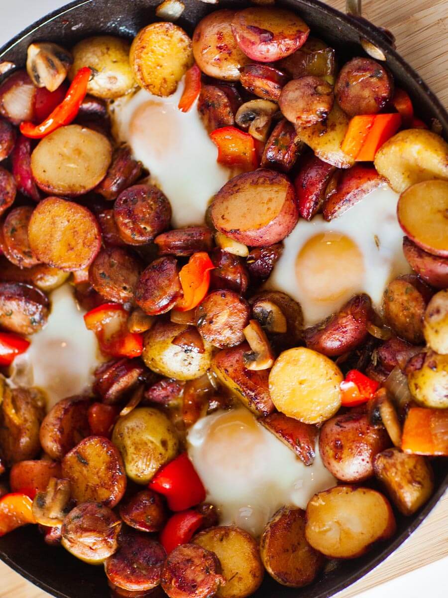 Close-up of camping breakfast skillet with a broken egg yolk running into crispy potatoes and sausage crumbles, garnished with fresh herbs and hot sauce on the side