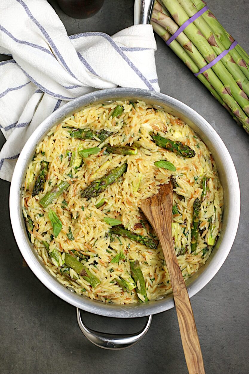 Ingredients for one pot cheesy orzo laid out on a marble surface — dry orzo, asparagus spears, wedge of Parmesan, garlic cloves, lemon, broth carton, and cream cheese, with a single wide pot nearby