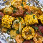 shrimp boil foil packets opened on a rustic wooden table with corn, sausage, shrimp and potatoes glistening in garlic butter