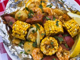 shrimp boil foil packets opened on a rustic wooden table with corn, sausage, shrimp and potatoes glistening in garlic butter