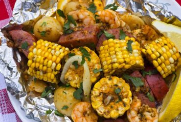 shrimp boil foil packets opened on a rustic wooden table with corn, sausage, shrimp and potatoes glistening in garlic butter