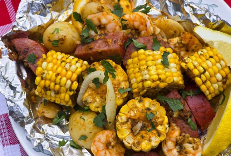 shrimp boil foil packets opened on a rustic wooden table with corn, sausage, shrimp and potatoes glistening in garlic butter