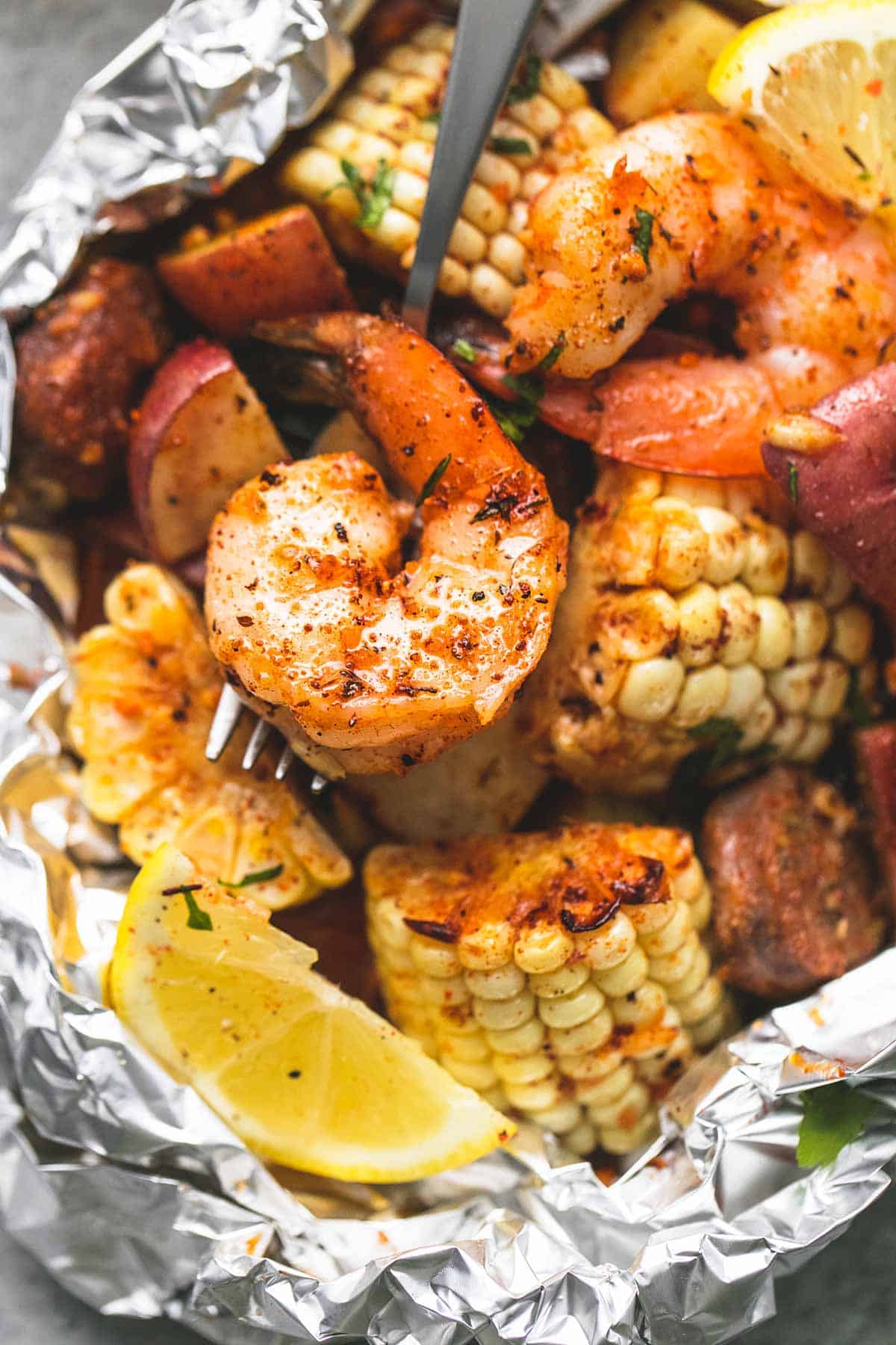 close-up of an opened shrimp boil foil packet with steam rising, showing glossy shrimp, charred corn, golden potatoes and sausage in garlic butter with a lemon wedge on the side