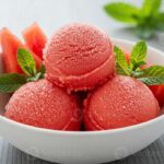 Scoops of vibrant pink-red watermelon sorbet served in a chilled white bowl garnished with fresh mint leaves and a wedge of watermelon on a sunny outdoor table