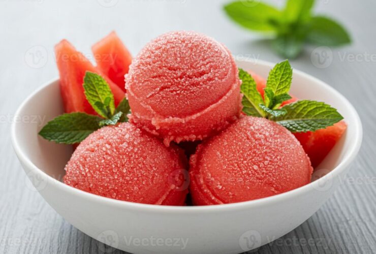 Scoops of vibrant pink-red watermelon sorbet served in a chilled white bowl garnished with fresh mint leaves and a wedge of watermelon on a sunny outdoor table