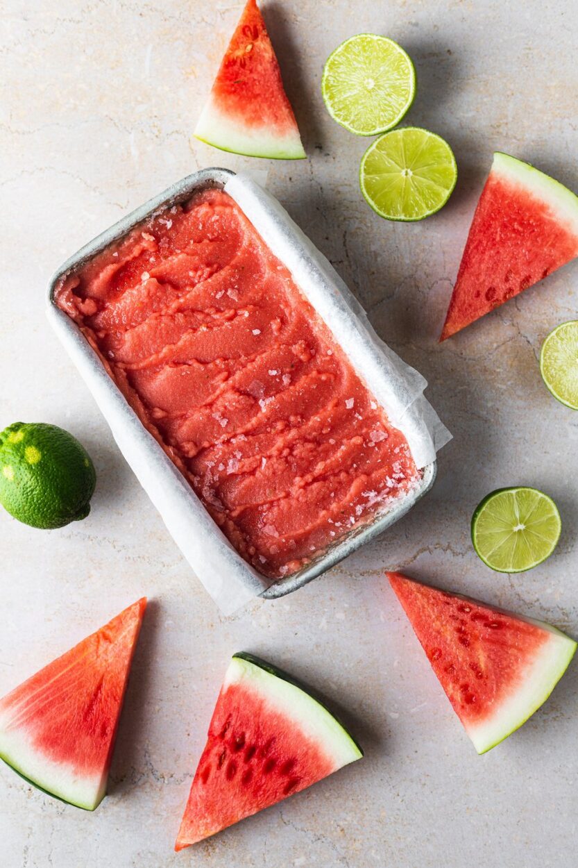 Overhead flat-lay of cubed watermelon chunks on a cutting board beside fresh limes, a small bowl of sugar, kosher salt, and a blender pitcher ready for prep