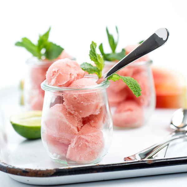 Close-up of a single scoop of watermelon sorbet in a vintage glass dessert cup, glistening with condensation, topped with a sprig of fresh mint and a thin lime wheel, photographed against a bright white background