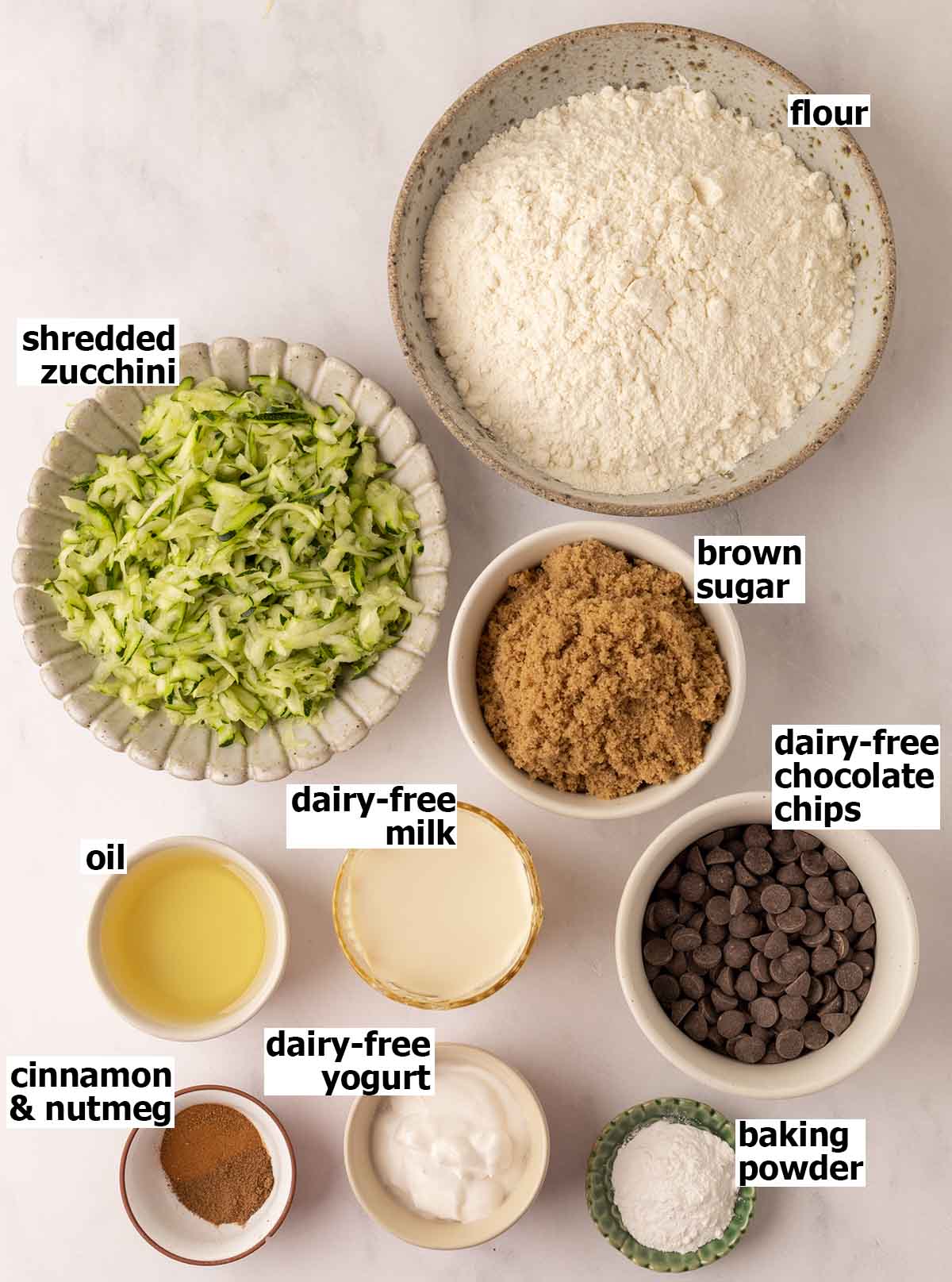 Overhead flat lay of zucchini bread ingredients — grated zucchini, eggs, brown sugar, cinnamon, oil, flour, and vanilla on a marble surface