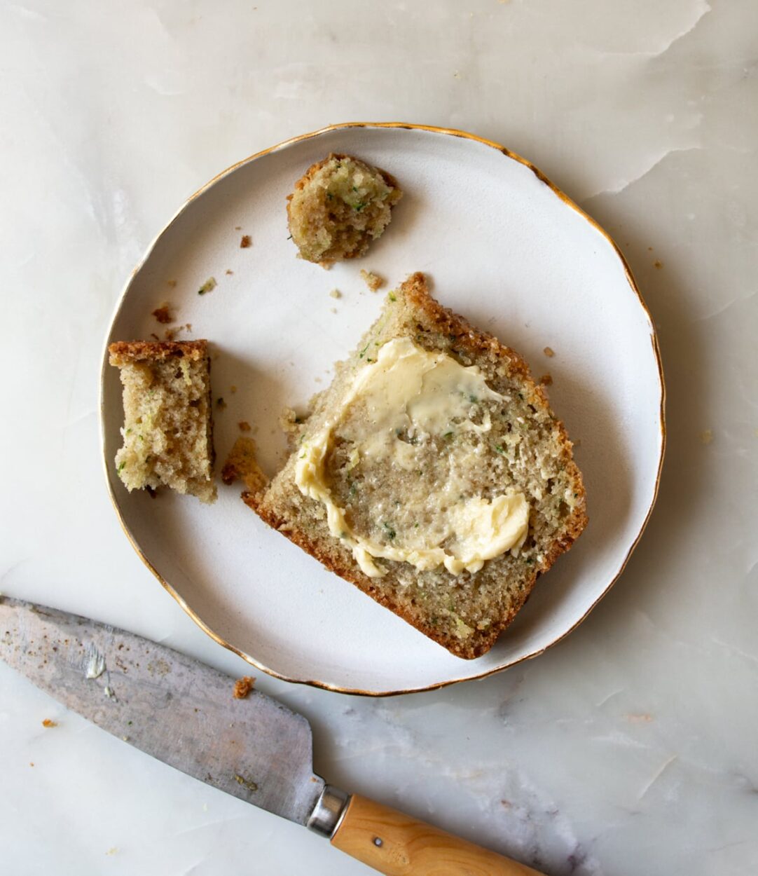 Close-up of a slice of zucchini bread showing moist golden crumb with cinnamon specks, served on a white plate with a pat of butter melting on top