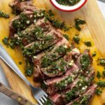 Argentinian chimichurri steak sliced and plated with vibrant green herb sauce drizzled over the top, served on a dark wooden board with grilled char marks visible