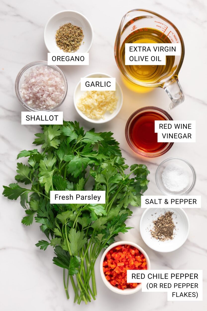 Overhead flat lay of chimichurri ingredients including fresh parsley, garlic cloves, red wine vinegar bottle, olive oil, red pepper flakes, and a bowl of chimichurri sauce in progress