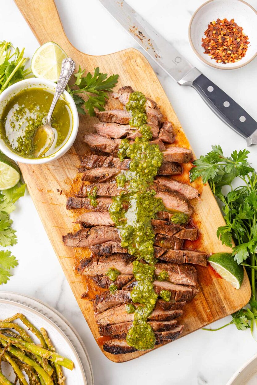 Close-up of sliced Argentinian chimichurri steak fanned out on a wooden board with chimichurri sauce spooned generously over the top, garnished with fresh parsley and a wedge of lemon