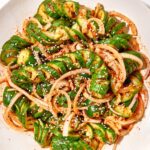 Asian cucumber noodle salad spiralized cucumber noodles in a white bowl with sesame seeds, sliced scallions, red chili, and golden sesame-ginger dressing, overhead shot on a wooden table