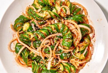 Asian cucumber noodle salad spiralized cucumber noodles in a white bowl with sesame seeds, sliced scallions, red chili, and golden sesame-ginger dressing, overhead shot on a wooden table
