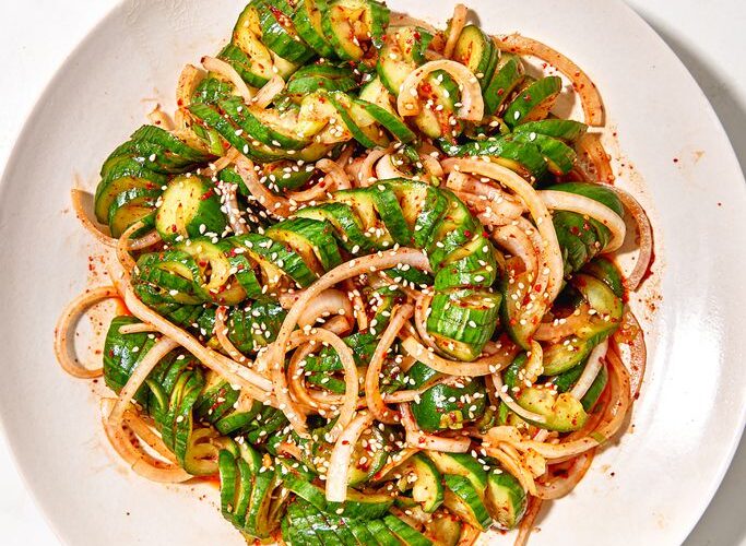 Asian cucumber noodle salad spiralized cucumber noodles in a white bowl with sesame seeds, sliced scallions, red chili, and golden sesame-ginger dressing, overhead shot on a wooden table