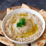 Baba Ganoush served in a wide shallow bowl drizzled with olive oil, topped with paprika, fresh parsley, and a side of warm pita bread on a rustic wooden table