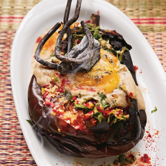 Whole eggplants being charred directly over a gas flame on a stovetop, skin blistering and blackening, with tahini, lemon, garlic, and olive oil arranged nearby on a marble surface