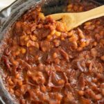 A cast iron skillet overflowing with thick, glossy BBQ baked beans topped with crispy bacon crumbles, fresh parsley, and a sticky caramelized sauce, photographed on a rustic wooden picnic table with a checkered napkin and a July 4th backdrop
