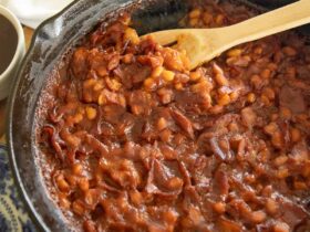 A cast iron skillet overflowing with thick, glossy BBQ baked beans topped with crispy bacon crumbles, fresh parsley, and a sticky caramelized sauce, photographed on a rustic wooden picnic table with a checkered napkin and a July 4th backdrop