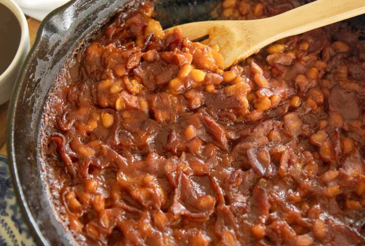 A cast iron skillet overflowing with thick, glossy BBQ baked beans topped with crispy bacon crumbles, fresh parsley, and a sticky caramelized sauce, photographed on a rustic wooden picnic table with a checkered napkin and a July 4th backdrop