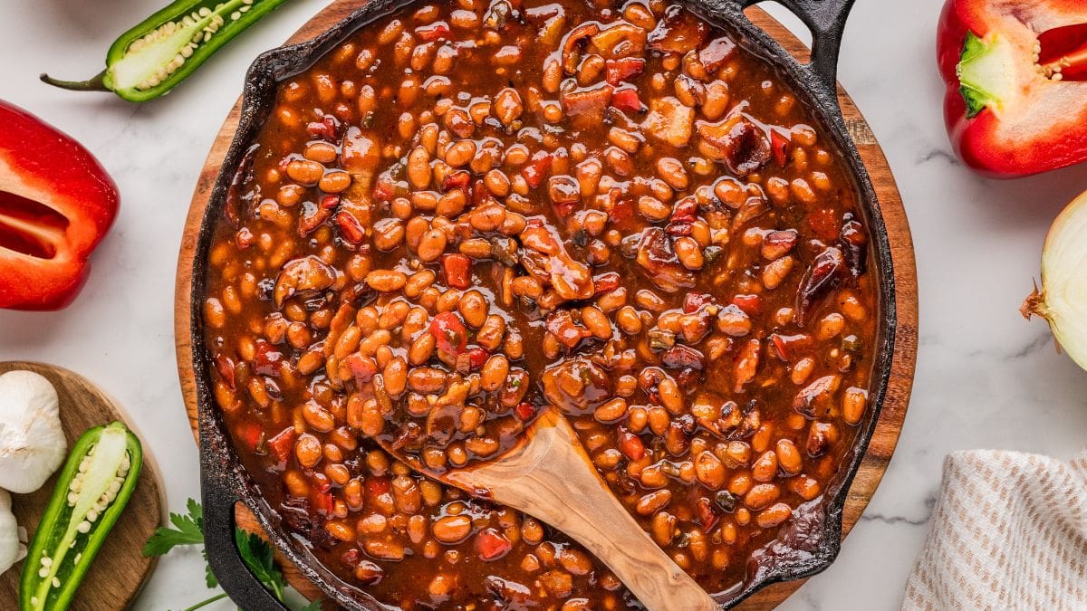 A close-up spoon lifting a heaping serving of thick, glossy BBQ baked beans with visible bacon chunks and caramelized edges, steam rising against a soft bokeh background of a summer cookout