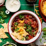 A bowl of thick beef chili topped with shredded cheddar cheese, sour cream, sliced jalapeños, and chopped green onions, photographed from above on a dark slate surface with a spoon resting beside it