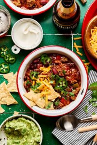 A bowl of thick beef chili topped with shredded cheddar cheese, sour cream, sliced jalapeños, and chopped green onions, photographed from above on a dark slate surface with a spoon resting beside it