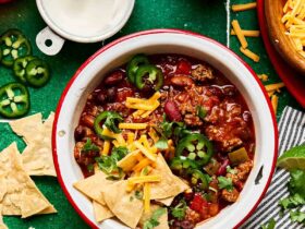 A bowl of thick beef chili topped with shredded cheddar cheese, sour cream, sliced jalapeños, and chopped green onions, photographed from above on a dark slate surface with a spoon resting beside it