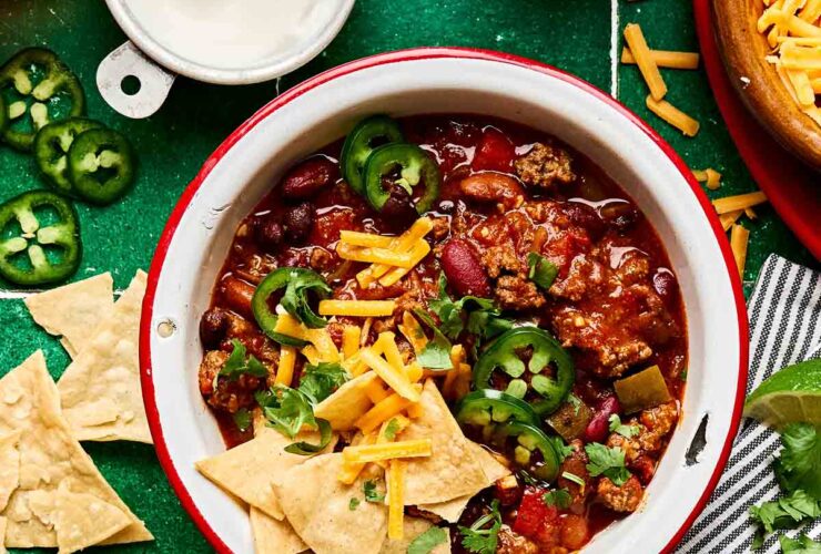 A bowl of thick beef chili topped with shredded cheddar cheese, sour cream, sliced jalapeños, and chopped green onions, photographed from above on a dark slate surface with a spoon resting beside it