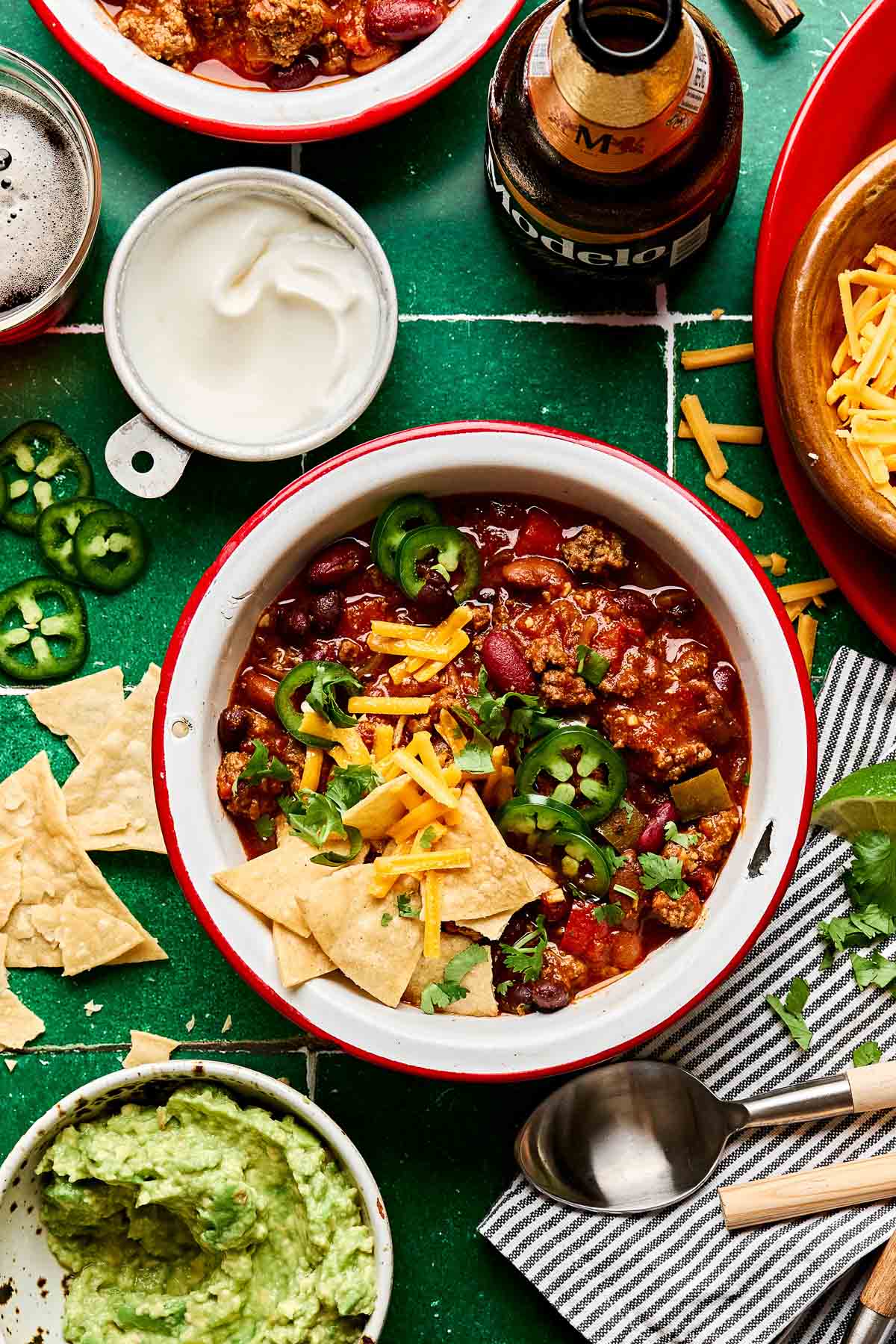 A bowl of thick beef chili topped with shredded cheddar cheese, sour cream, sliced jalapeños, and chopped green onions, photographed from above on a dark slate surface with a spoon resting beside it